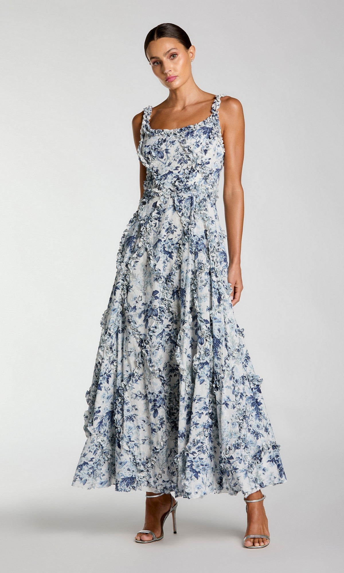 A woman wears the Mac Duggal 8256 Floral Wedding Guest Dress in Blue Multi, a long sleeveless floral print with ruffled details and an a-line skirt, paired with high-heeled sandals against a plain background.